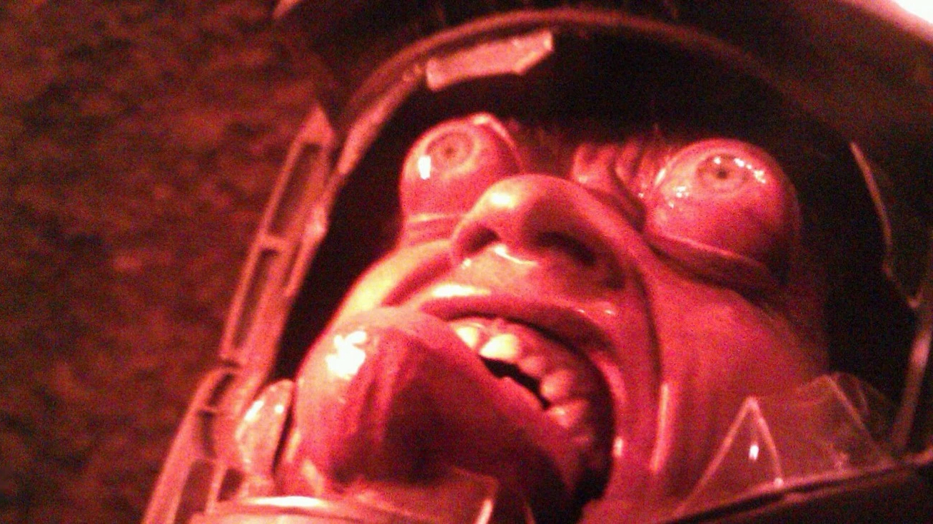 a man's eyes and tongue bulge from his face inside a cracked spacesuit helmet