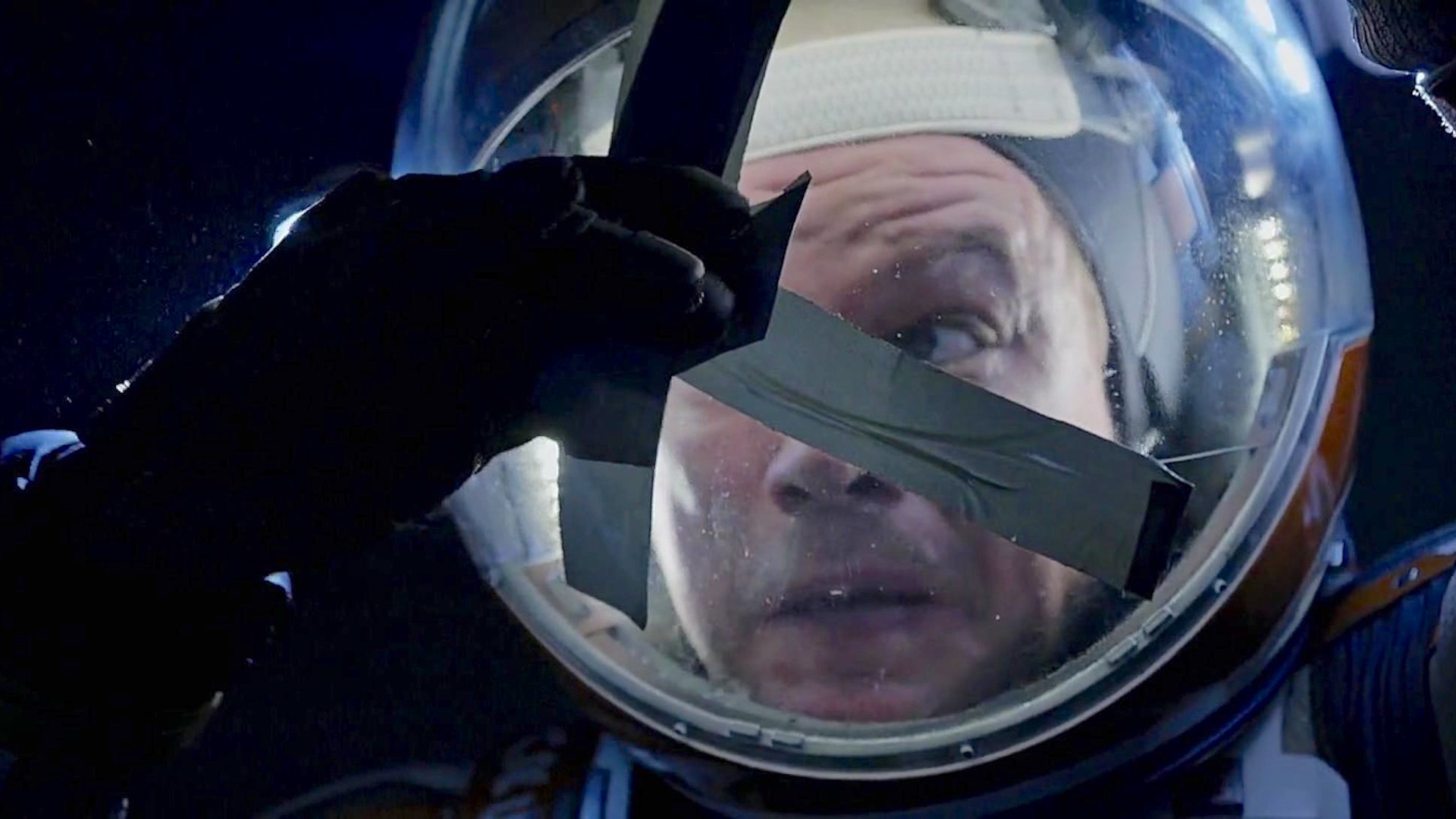 a man puts duct tape over a crack in his spacesuit's glass visor
